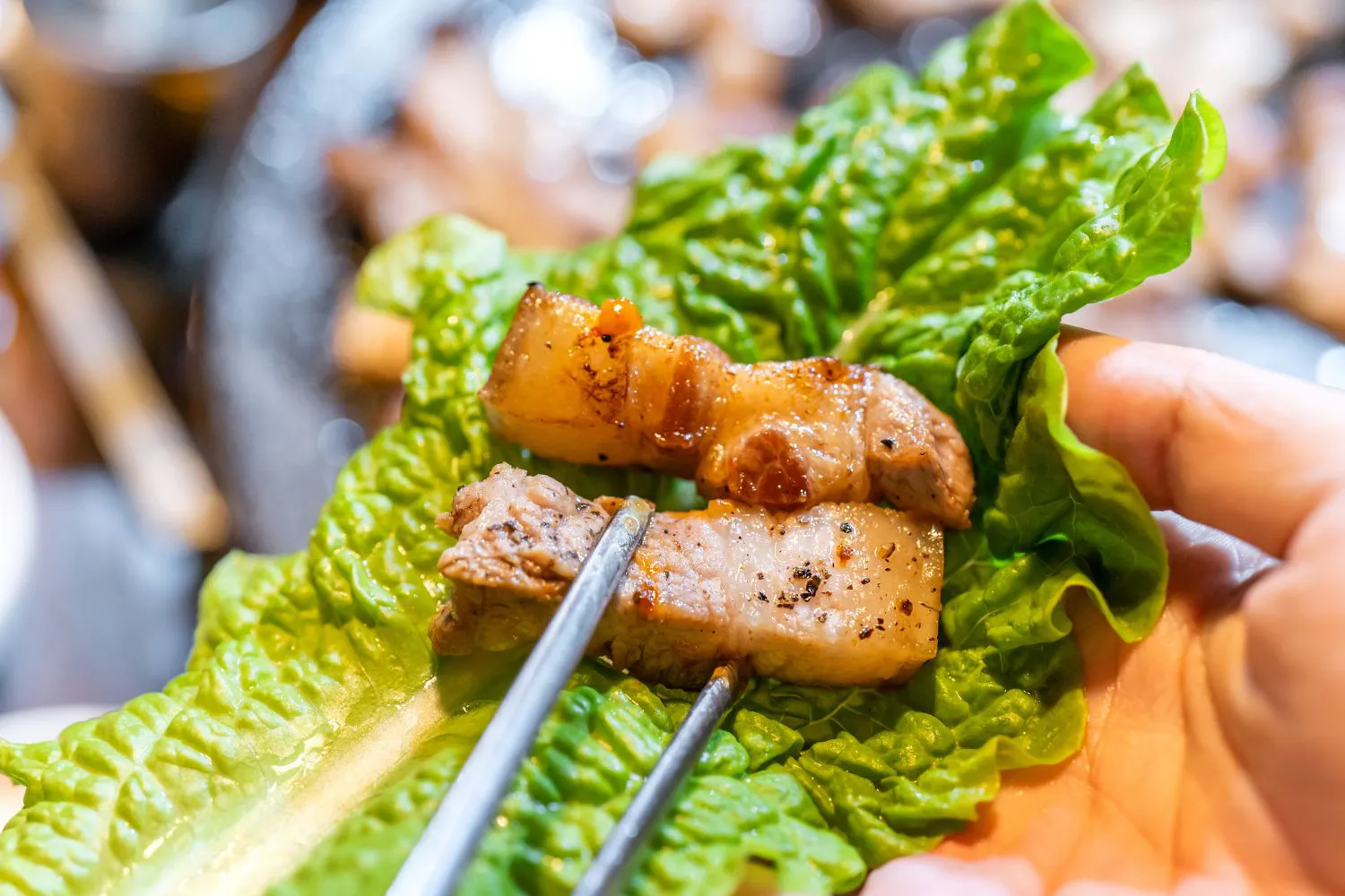 Two pieces of grilled pork belly resting on lettuce held with metal tongs at Feast Utopia Clackamas, an Asian Fusion Restaurant in Clackamas