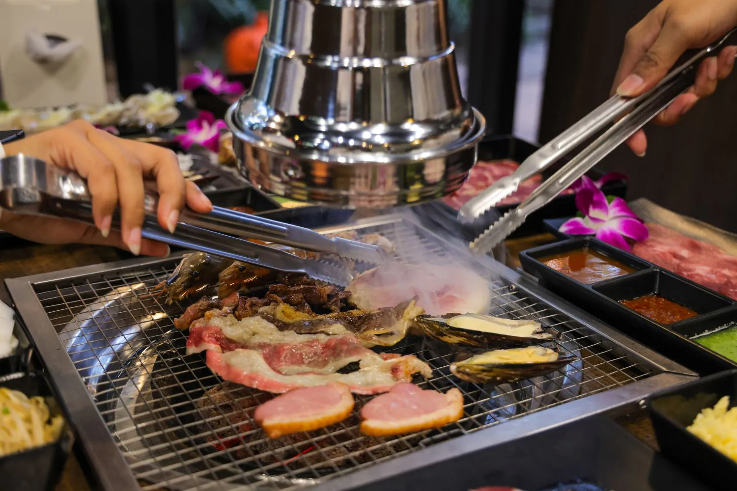 Two hands using metal tongs to grill mixed meat, shrimp and mussels on Korean BBQ mesh at Feast Utopia Clackamas, an Asian Fusion Restaurant in Clackamas